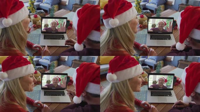 微笑的高加索夫妇戴着圣诞老人的帽子，使用笔记本电脑与屏幕上的男人进行圣诞节视频通话