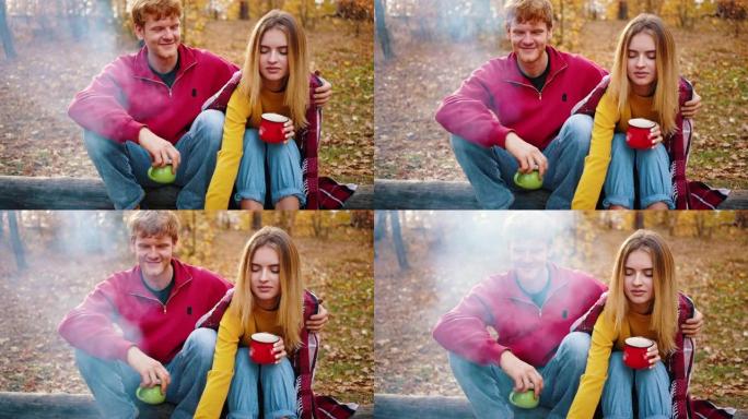 年轻人拥抱女友，女友坐在秋天的木头上时捧着一杯茶。野餐，篝火。特写，慢动作