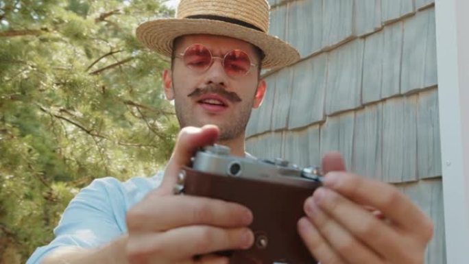 老式花哨的年轻摄影师时髦的男人，戴着草帽的小胡子，粉色太阳镜摆姿势，在乡村别墅的旧电影相机上自拍。现