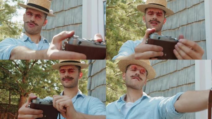 老式花哨的年轻摄影师时髦的男人，戴着草帽的小胡子，粉色太阳镜摆姿势，在乡村别墅的旧电影相机上自拍。现