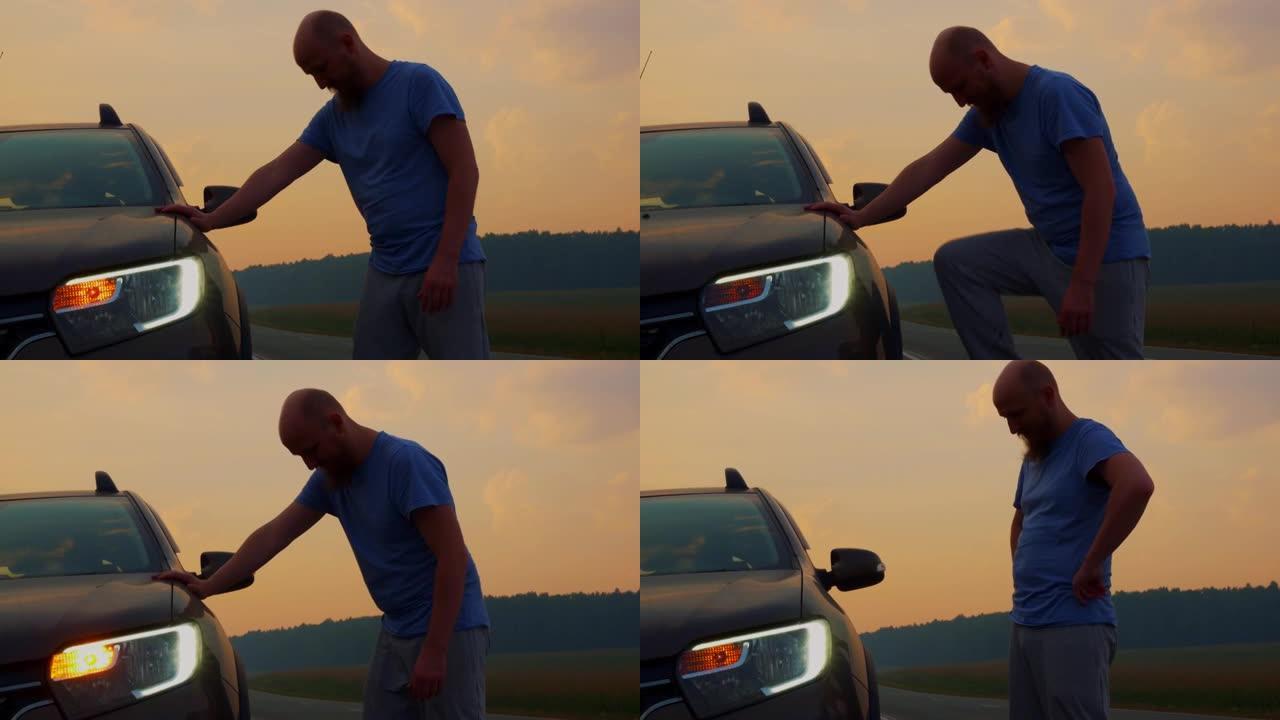一个秃头的大胡子男人站在路边的破车旁。路上的麻烦。美丽的夕阳照亮了汽车，大灯和警示灯亮起