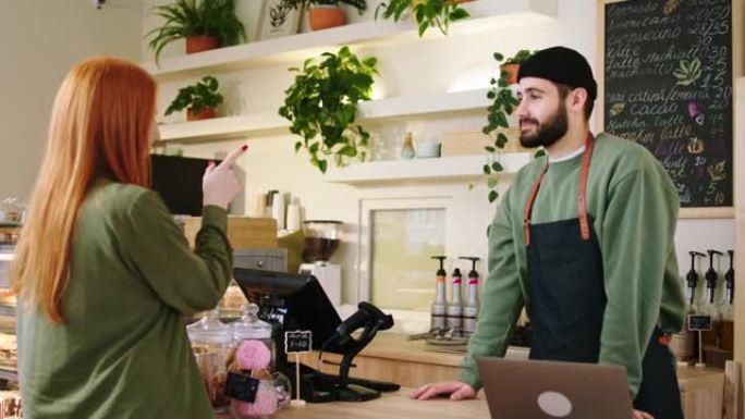 在拥有完美氛围的现代咖啡店中，魅力四射的咖啡师使用智能手机从客户那里获得在线订单并在笔记本电脑上打字