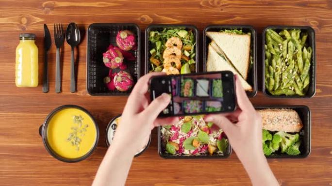 送餐俯视图，在木制桌子上的一次性容器中带走餐点。在智能手机上拍摄带有煮熟的素食菜肴的午餐盒照片。健康