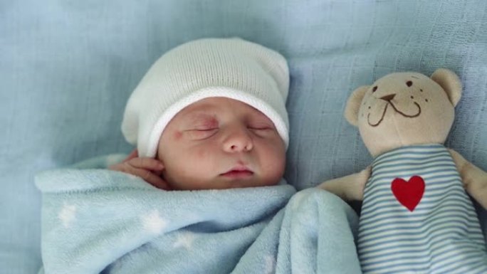 初生婴儿脸部肖像的特写镜头早期与蓝色背景上的塔迪熊睡觉。孩子在帽子上生命的开始。婴儿，分娩，出生的最
