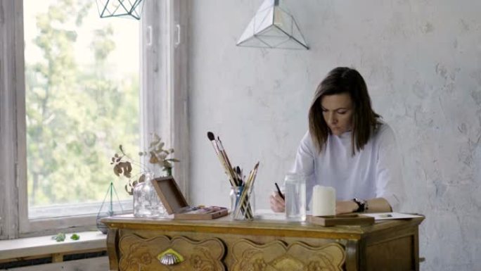 一位漂亮的黑暗女性艺术家在木桌上用铅笔在纸上画画。4K