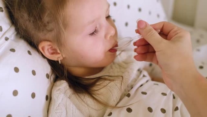 两岁的生病的小孩躺在床上，喝妈妈给的勺子里的药。母亲照顾一个生病的孩子。药用药水和退热药物。新型冠状