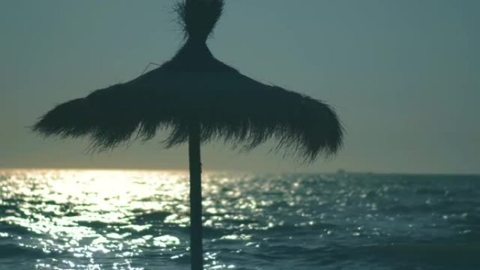 草伞在夏天的微风中挥舞着，海面上有地平线，日落的光线反射在水面上。热带海滩和暑假概念