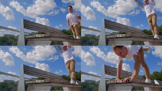 在城市背景下，杂技演员在玻璃人行天桥的长凳上表演跑酷技巧的前视图特写镜头。穿着米色裤子和白色t恤的高
