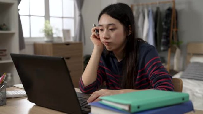 无聊的亚洲女学生在家学习正在玩笔和支撑脸，同时在计算机上观看无聊的在线视频讲座