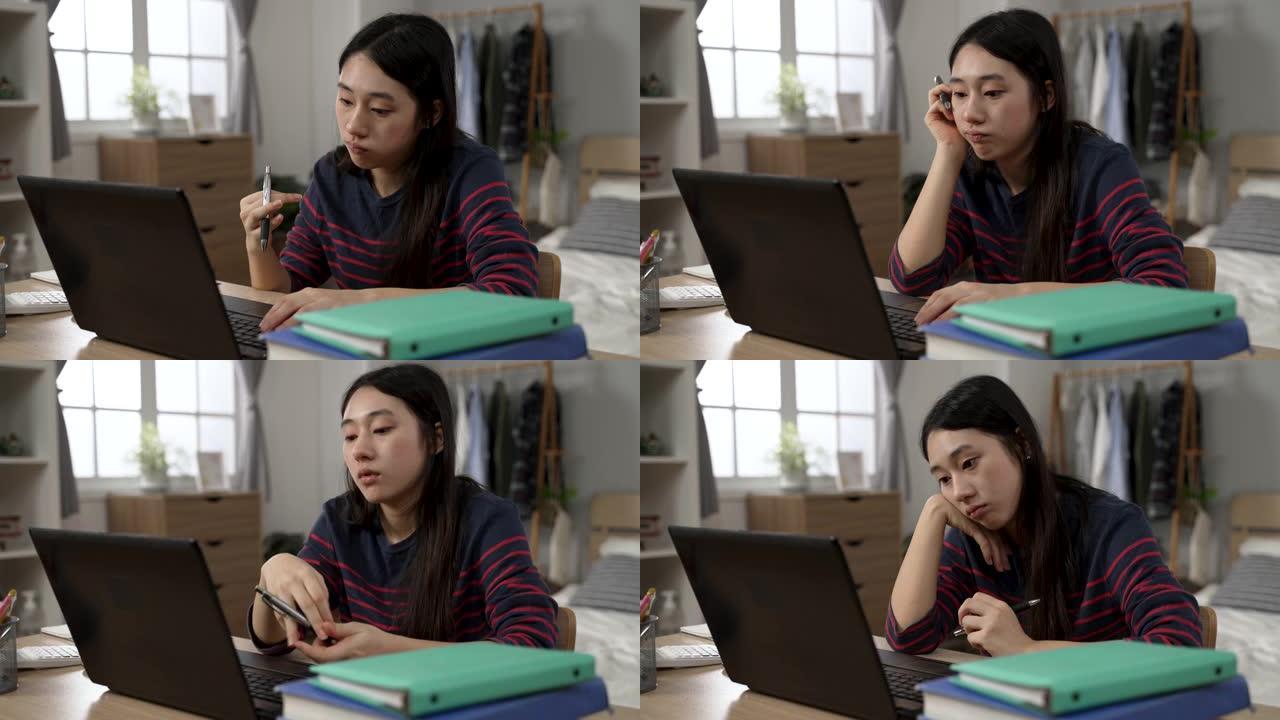 无聊的亚洲女学生在家学习正在玩笔和支撑脸，同时在计算机上观看无聊的在线视频讲座