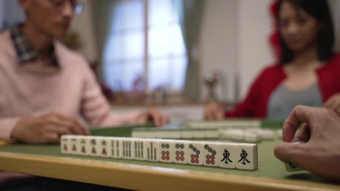 特写与选择性焦点的一个玩家的手玩瓷砖旁边的麻将棋子的手，而他的家人正在有他们的回合。中文翻译:东方