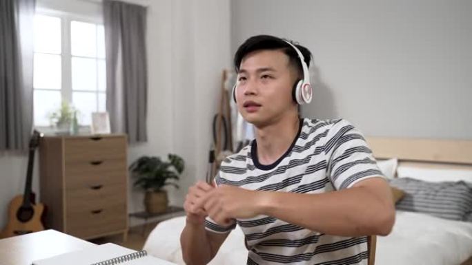 分心的亚洲年轻人从家里学习，正在玩笔像鼓棒，同时在卧室的办公桌上听耳机上的流行歌曲。