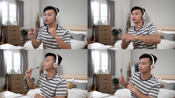 分心的亚洲年轻人从家里学习，正在玩笔像鼓棒，同时在卧室的办公桌上听耳机上的流行歌曲。