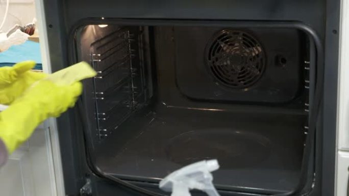 妇女在厨房做饭后戴着橡胶手套清洁烤箱，用烤箱清洁剂喷洒并用布擦拭。