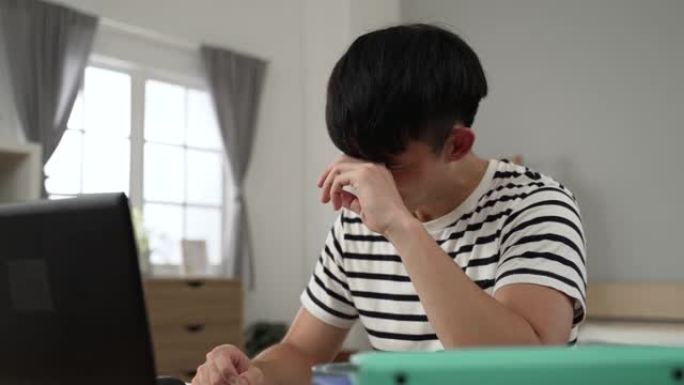 压力很大的亚洲男学生在卧室里检疫时，在家里看笔记本电脑上的视频讲座时揉眼睛