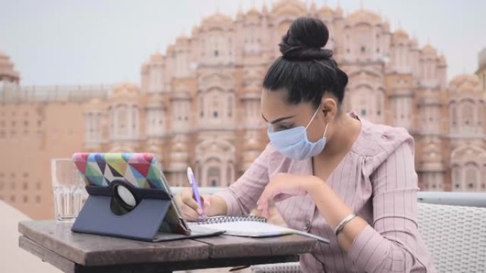 一位迷人的现代女性正坐在一家咖啡馆里，戴着面部防护面具，用数码平板电脑进行在线视频通话