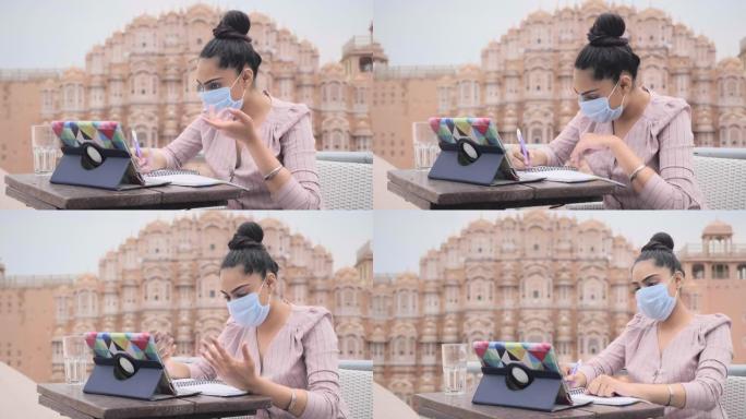一位迷人的现代女性正坐在一家咖啡馆里，戴着面部防护面具，用数码平板电脑进行在线视频通话