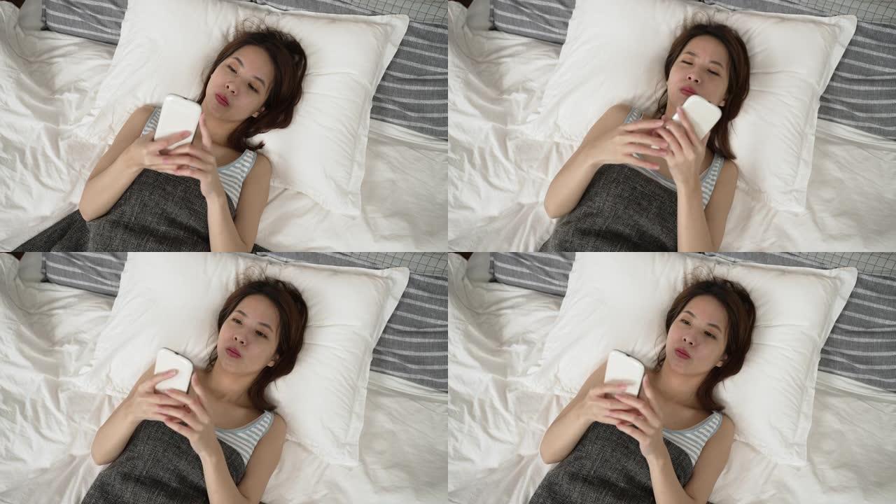 亚洲女性躺在床上玩智能手机。粗心的女孩在卧室放松，在手机上打字。心不在焉的女人看着手机，早上摔倒在胸