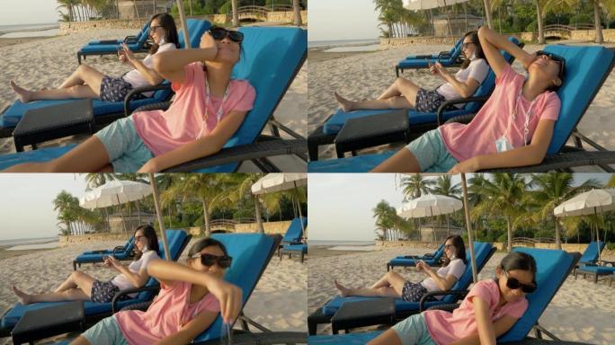 快乐的亚洲女孩在沙滩椅上放松，母亲在度假期间一起在酒店或度假村使用智能手机，生活方式理念。
