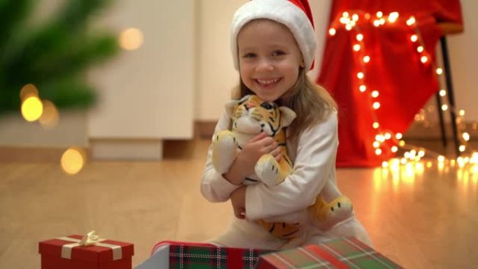 戴着红色圣诞老人帽子的小女孩坐着打开的盒子和玩具老虎，看着相机，在圣诞树旁边微笑。新年的象征