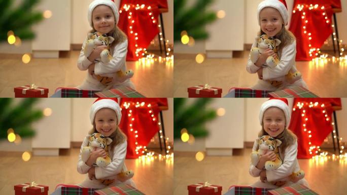戴着红色圣诞老人帽子的小女孩坐着打开的盒子和玩具老虎，看着相机，在圣诞树旁边微笑。新年的象征