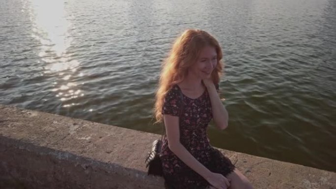 穿着夏装的红发女士坐在河边风化的混凝土栏杆上，她开心地微笑着