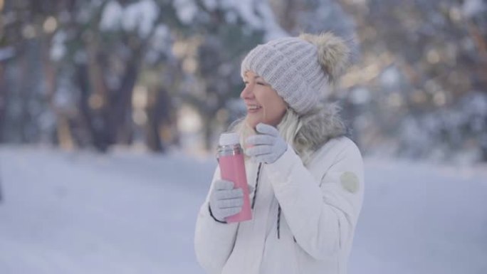 穿着温暖的冬装可爱的女孩的肖像从热水瓶里喝热茶。年轻女子在白雪皑皑的森林模糊背景下在户外摆姿势。特写