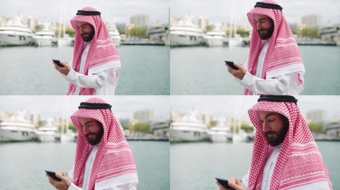 传统着装的阿拉伯商人在港口使用智能手机，在手机上阅读互联网新闻