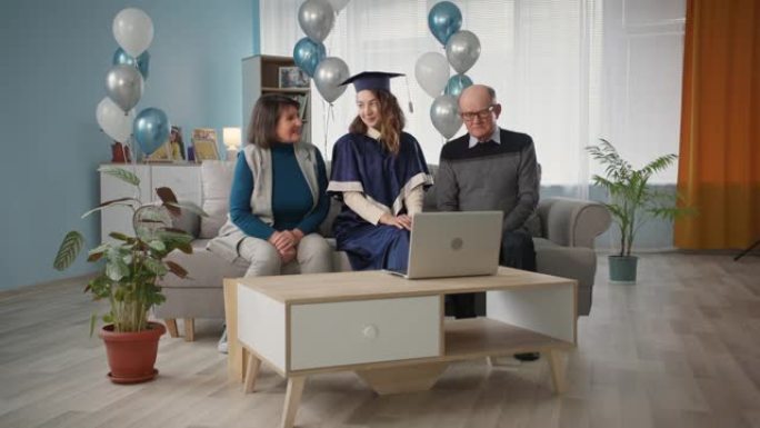 毕业，带着女儿穿着学术服装的快乐家庭在家坐在沙发上庆祝网上毕业典礼气球背景关于社交距离时间