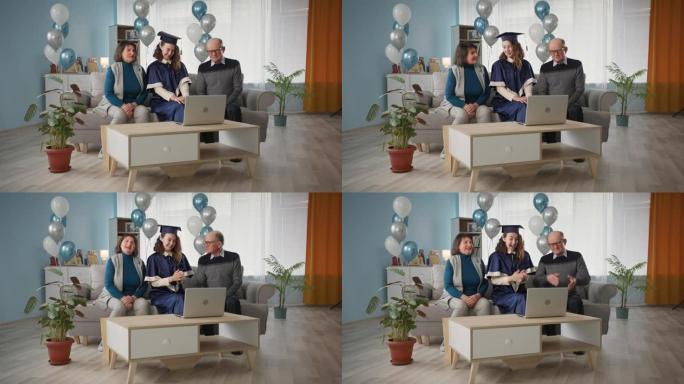 毕业，带着女儿穿着学术服装的快乐家庭在家坐在沙发上庆祝网上毕业典礼气球背景关于社交距离时间