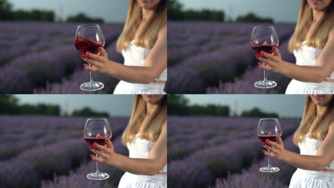 在薰衣草田的背景下，一个女孩的手和一杯红酒的特写镜头。年轻的金发酒瘾淡紫色灌木丛。喝薰衣草。