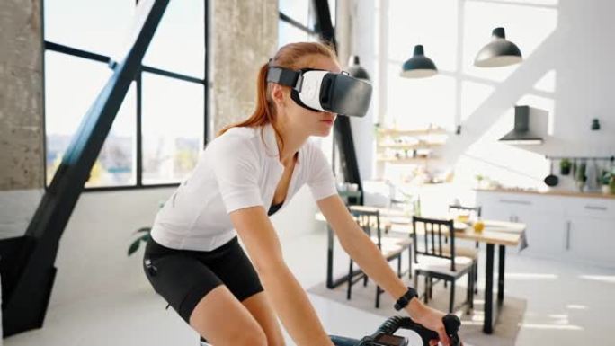 VR耳机中的年轻女子正在踩踏室内自行车教练。工作室公寓，厨房。在家进行日常训练。特写，慢动作