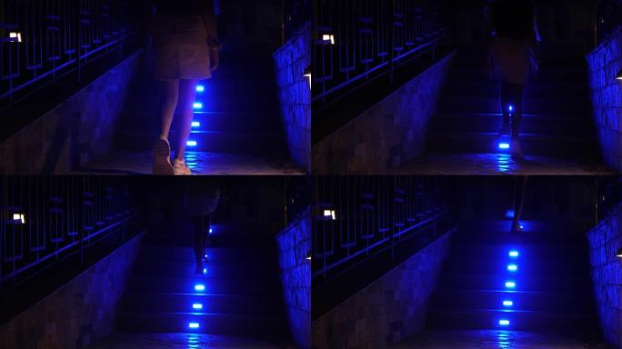 照明楼梯台阶。特写。女性腿部的轮廓，穿着运动鞋，爬上台阶，明亮的蓝色照明，晚上，在酒店大楼的花园中。