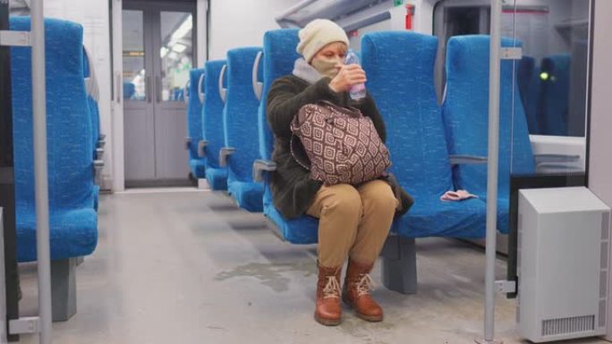 戴着面部保护面具的成熟女人正在乘坐旅客列车。她正在喝瓶装水。