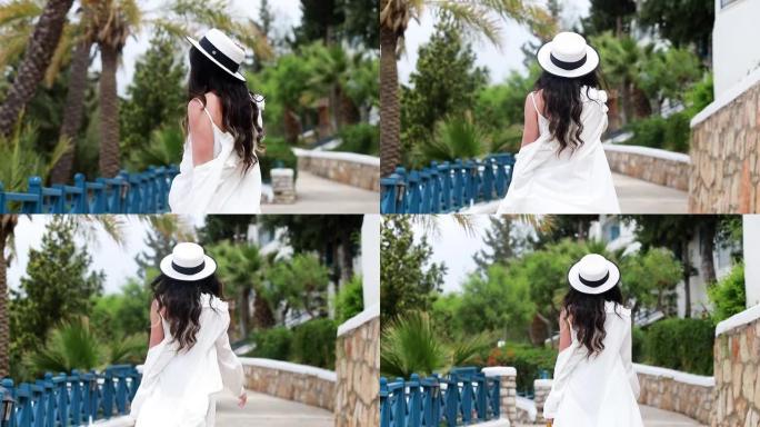 年轻微笑的女人穿着白色连衣裙，手里戴着草帽走路