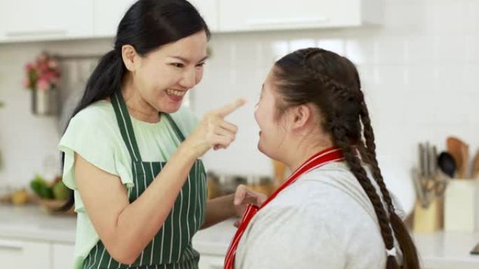 开朗的亚洲母亲在厨房一起做饭并玩得开心时，在患有唐氏综合症的可爱女孩的鼻子上涂抹面粉