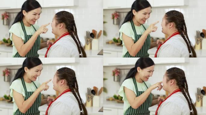 开朗的亚洲母亲在厨房一起做饭并玩得开心时，在患有唐氏综合症的可爱女孩的鼻子上涂抹面粉