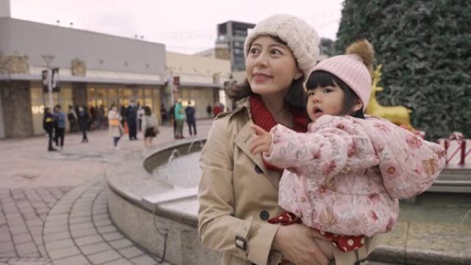 亚洲母亲和她的小女儿在一个有圣诞装饰品的城市广场上度过快乐的时光。可爱的女孩好奇地指着远方