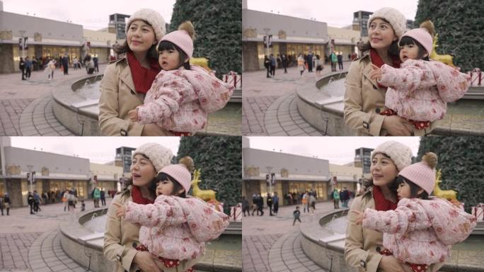 亚洲母亲和她的小女儿在一个有圣诞装饰品的城市广场上度过快乐的时光。可爱的女孩好奇地指着远方