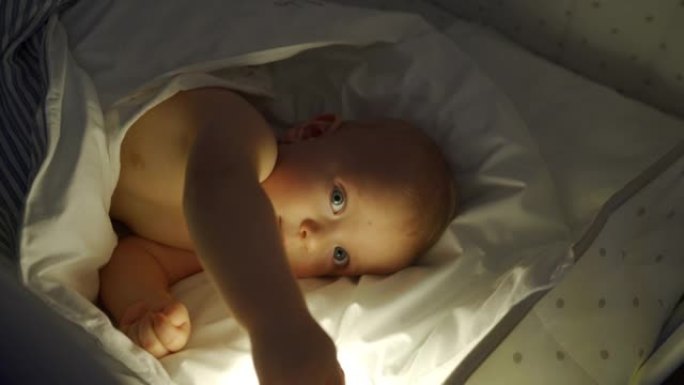 晚上，在夜灯的灯光下，将9个月的男婴包裹在婴儿床中。快乐婴儿从襁褓中挣脱出来的脸。