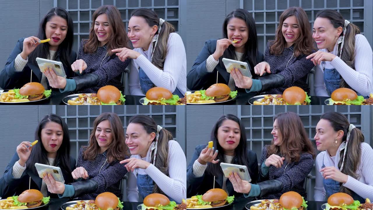 三名年轻女子坐在餐桌旁吃汉堡和薯条，看着智能手机微笑。