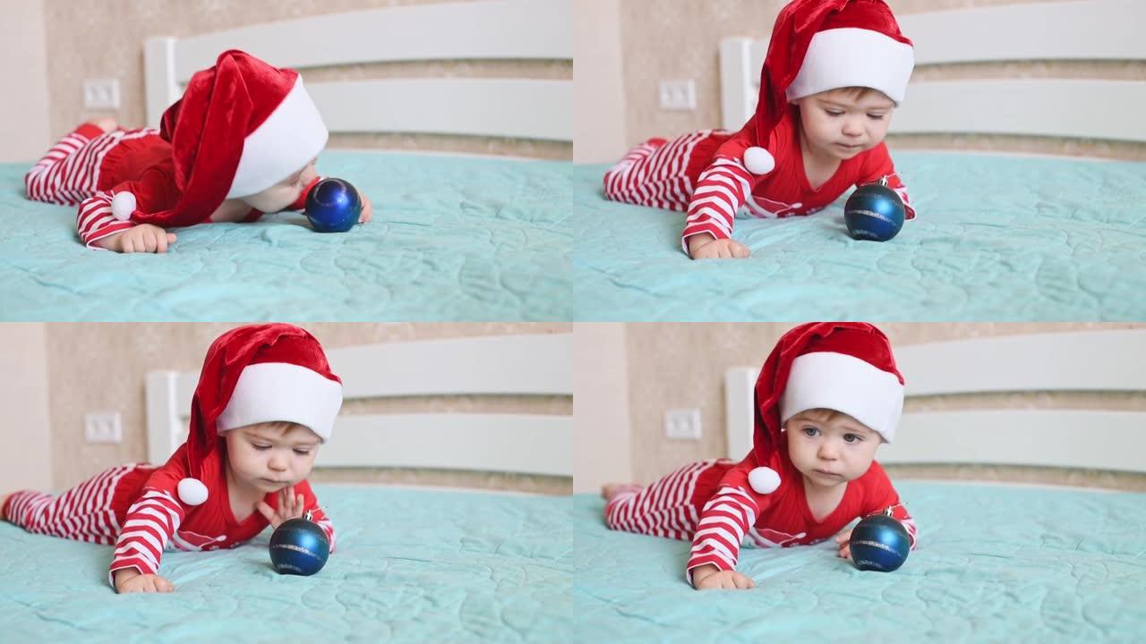 一个戴着圣诞老人帽子的一岁婴儿躺在肚子上，玩猩红色的圣诞球。穿着红色白色条纹西装的孩子。圣诞快乐，新