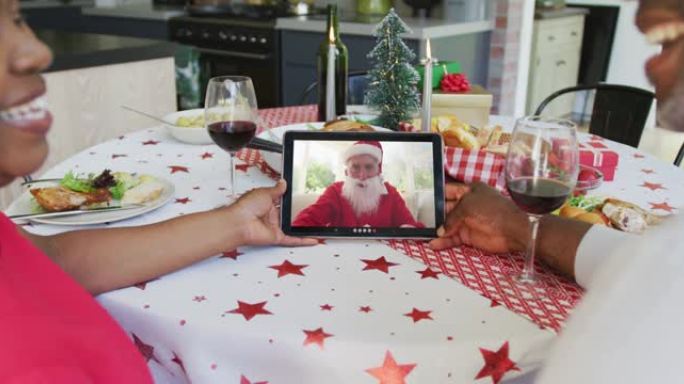 非裔美国人夫妇与葡萄酒使用平板电脑进行圣诞节视频通话，屏幕上有快乐的圣诞老人