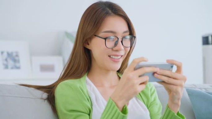 亚洲美女戴眼镜，在家里用手机聊天。迷人的年轻女孩坐在沙发上，使用智能手机后感到兴奋和快乐在客厅在线查