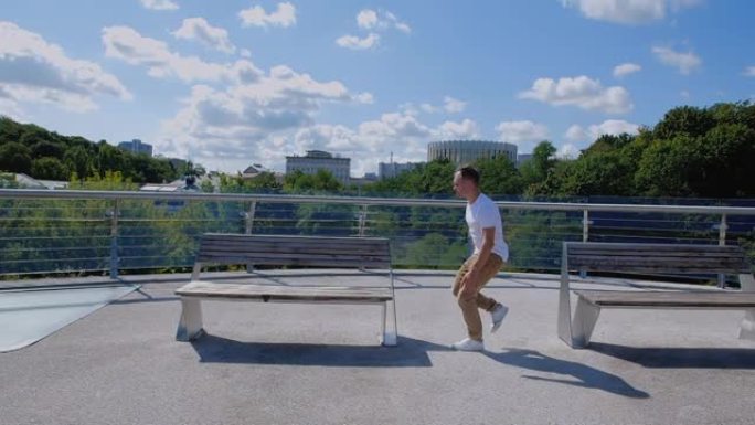 在城市背景下，杂技演员在玻璃人行天桥的长凳上表演跑酷技巧的跟踪镜头。穿着米色裤子和白色t恤的高加索人