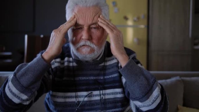 老人按摩头部，看着相机。头痛和头部健康问题。患有偏头痛的老人