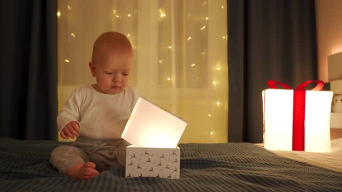 可爱的男婴拿着圣诞或生日礼物，高加索孩子在卧室的床上打开礼品盒，配有节日照明。生日，圣诞节，新年庆祝