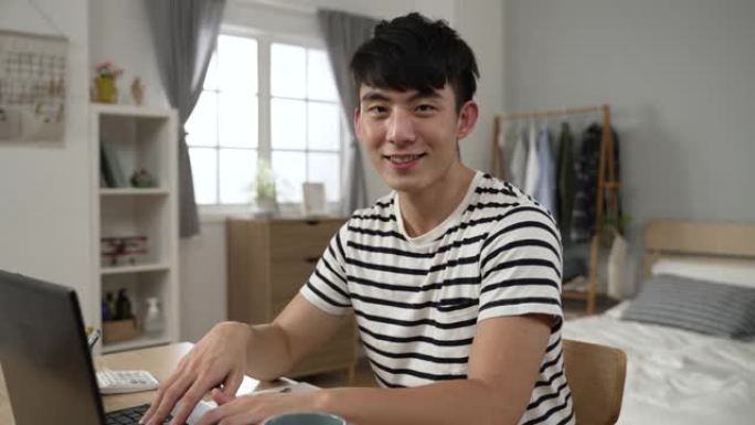一名亚洲男成年学生在锁定期间用笔记本电脑在家学习时微笑着看着相机的肖像