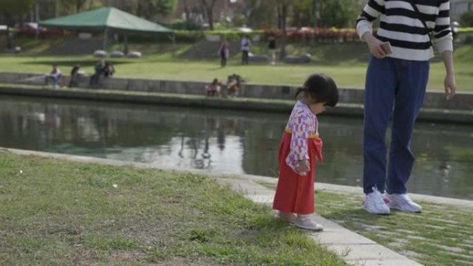 一名身着传统服饰的日本幼童在公园池塘边和妈妈玩耍，好奇地向下看着草坪