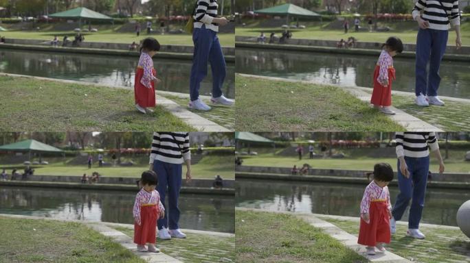 一名身着传统服饰的日本幼童在公园池塘边和妈妈玩耍，好奇地向下看着草坪